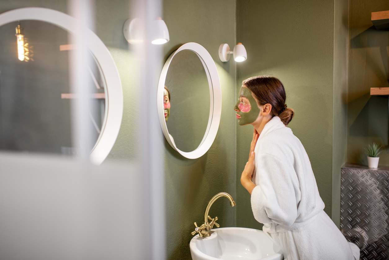 Woman in bathrobe taking care of herself with facial mask looking into the mirror in the beautiful green bathroom