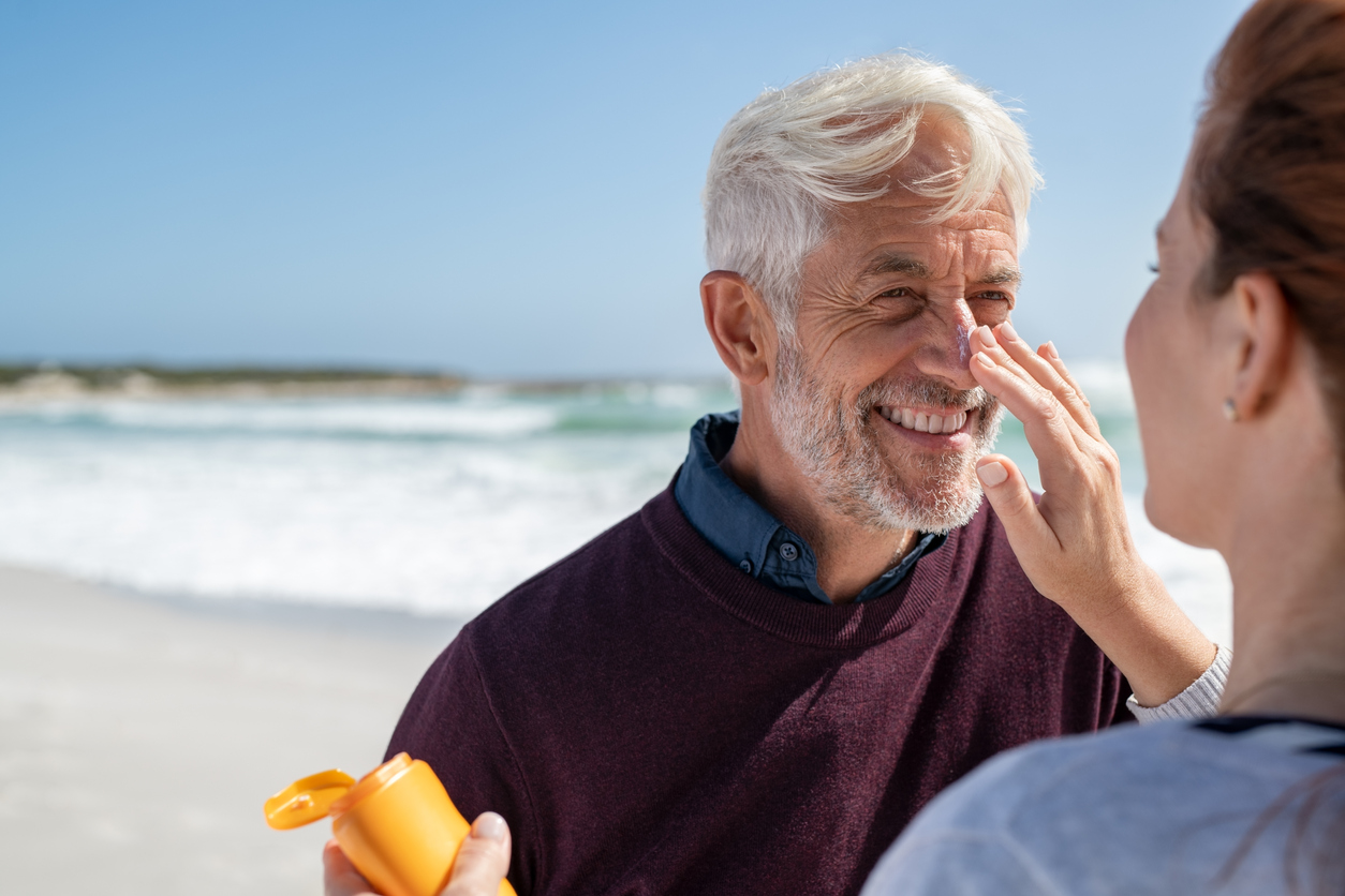 Portrait of cheerful old man looking at his mature wife applying sunscreen on nose. Senior husband enjoying vacation with woman while applying sunscreen on face at beach. Middle aged retired couple applying suntan lotion at sea with copy space.