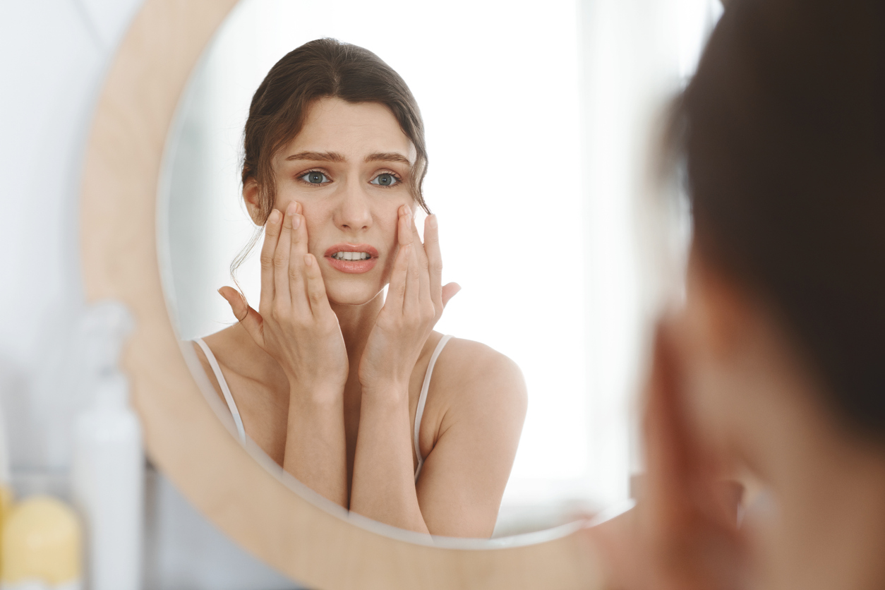 First wrinkles. Stressed girl touching her skin and looking at mirror at home