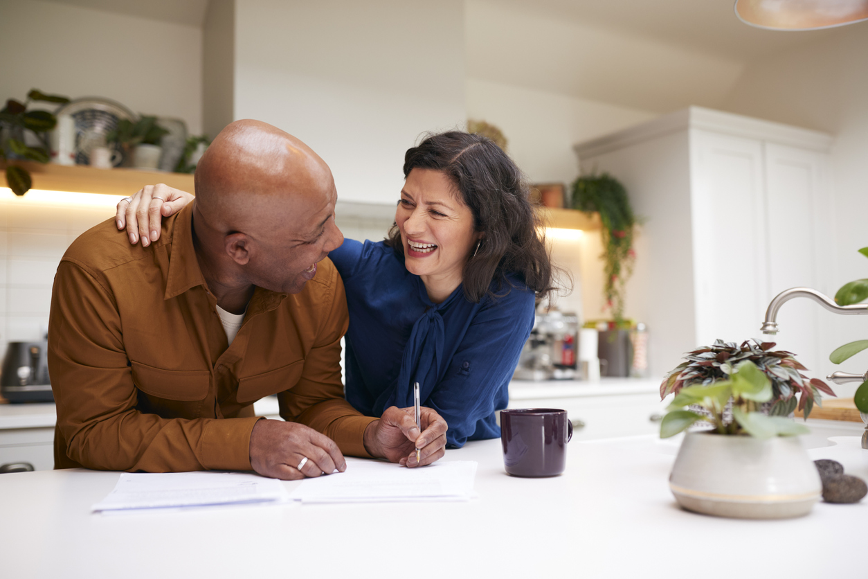 Mature Couple Reviewing And Signing Domestic Finances And Investment Paperwork In Kitchen At Home