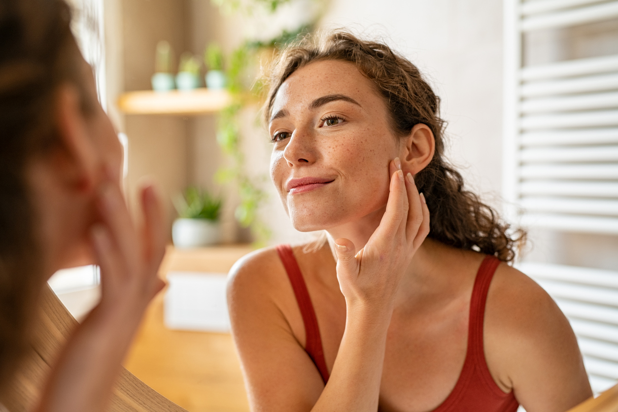 Beauty girl looking at mirror while touching her face and checking pimple, wrinkles and bags under the eyes, during morning beauty routine. Smiling pretty woman applying cream on her face in bathroom. Happy smiling beautiful young woman applying moisturizer.